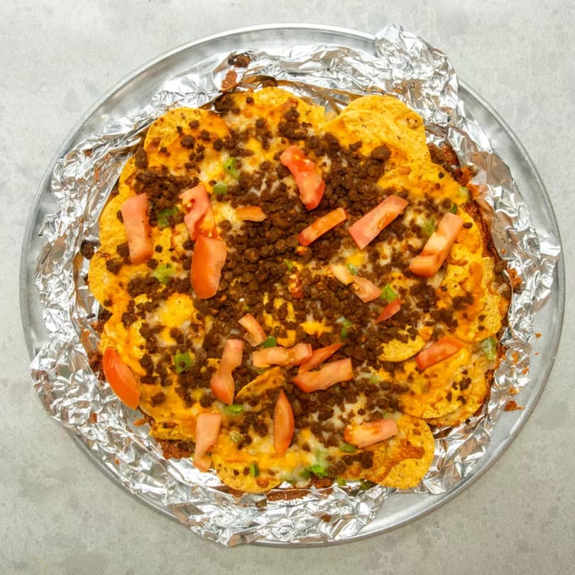 A round pan of nachos with melted cheese, ground beef, diced tomatoes, and green onions.
