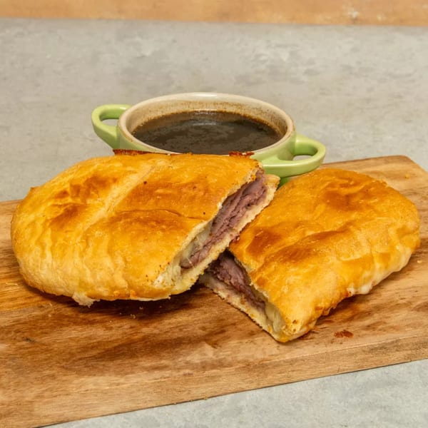 Golden-brown baked sandwich, sliced in half, revealing roast beef and cheese, with a side of au jus.