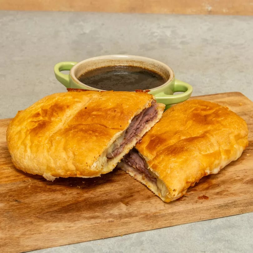 Golden-brown baked sandwich, sliced in half, revealing roast beef and cheese, with a side of au jus.