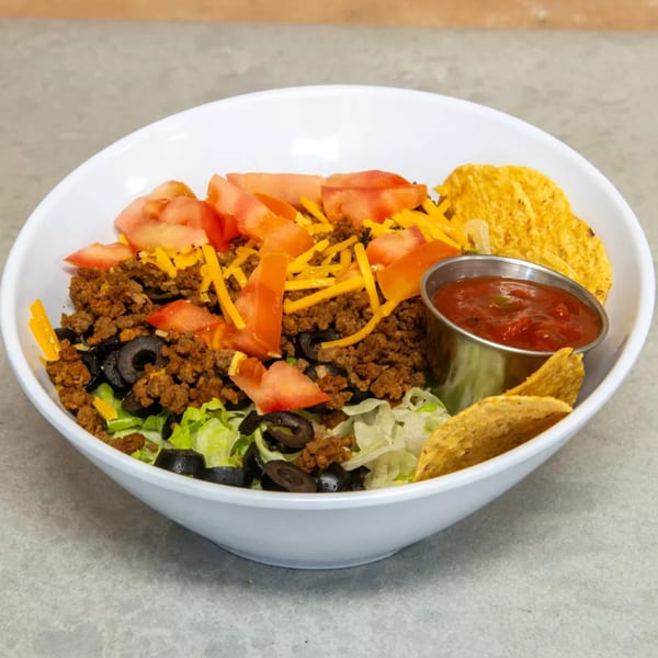 A taco salad in a white bowl with ground beef, lettuce, tomatoes, olives, cheese, and salsa.