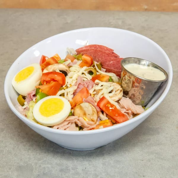 A white bowl filled with a chef salad, hard-boiled eggs, salami, and a side of dressing.