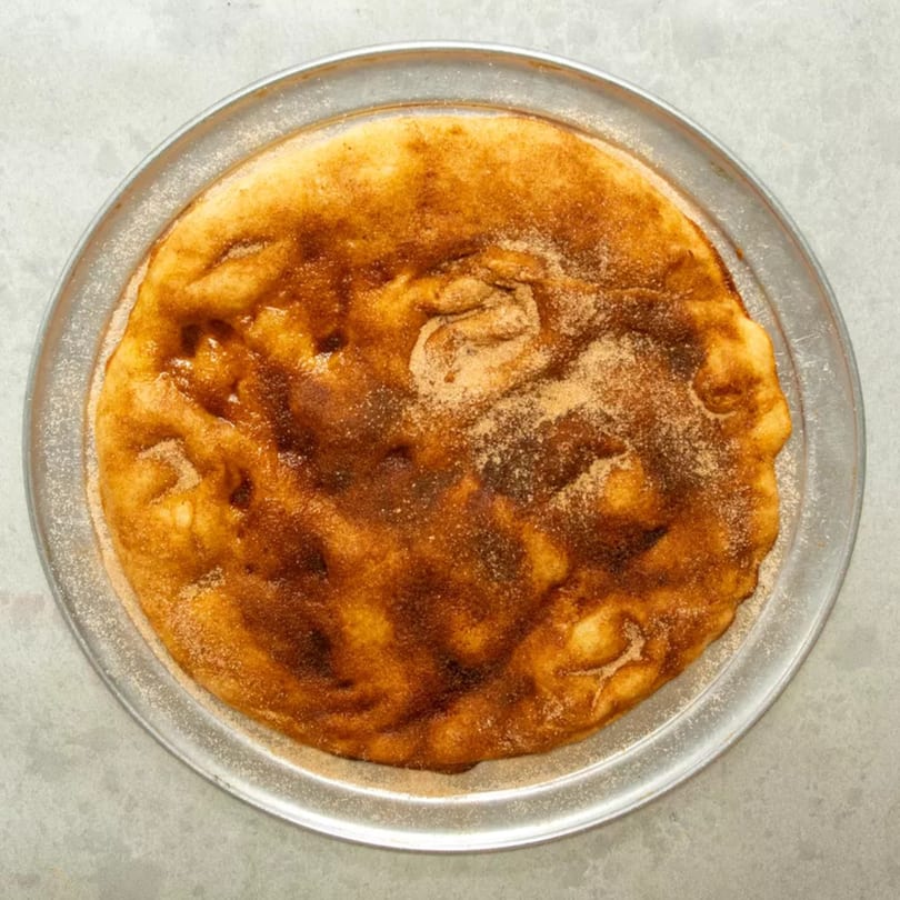 A golden-brown dessert pizza, sprinkled with cinnamon sugar, rests on a metal pan.